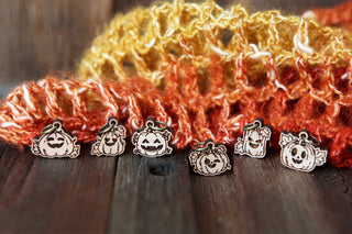 Six 🎃 Pumpkin Jack O’Lantern Stitch Markers with unique faces are arranged on a wooden surface in front of an orange and yellow knit scarf, set against a softly blurred wood background.