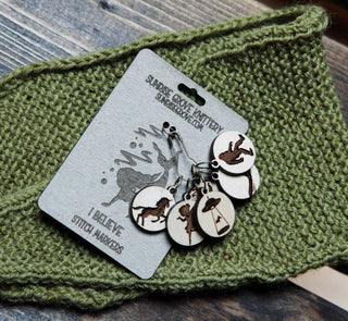 The 👽 I Believe Stitch Marker Set 👽 includes maple wood stitch markers with laser-etched cryptid designs, attached to a labeled card and shown on green knit fabric—ideal knitting accessories for any cryptid enthusiast.