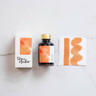 A bottle of Bottled Ink in Marmalade with a gold cap sits between its white box and a paper displaying vibrant orange strokes, all on a white surface—ideal for those who appreciate distinctive inks.