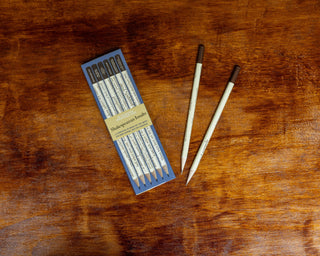 The Shakespearean Insults Pencil Set features six light-barreled pencils with brown erasers in a box, displayed on wood, accompanied by two extra literary pencils.
