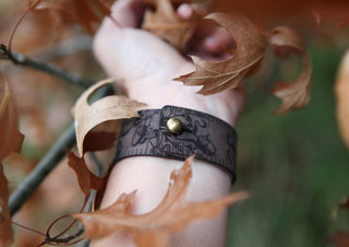 A close-up of a wrist wearing the Oak Leaves & Acorn Leather Ruler Cuff, crafted from reclaimed leather with a brass button, surrounded by dry autumn leaves on a tree branch.