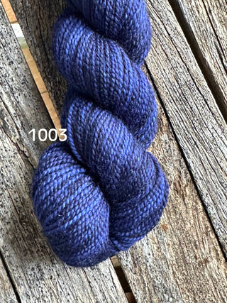 A skein of Koigu KPM & KPPPM Fingering Weight Yarn in deep blue (color 1003) rests on a weathered wooden surface. Ideal for knitting, crochet, or any cozy yarn project.