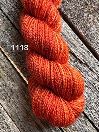 A skein from the KPM and KPPPM Variegated Color Collection in shade 1118—orange with subtle red tones—rests on weathered wood, perfect for your next hand knitting project.
