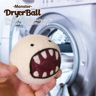 A hand holds a white Wool Felted Monster Dryer Ball with felted sharp teeth and a dark mouth in front of an open washing machine. Text reads: Wool Felted Monster Dryer Balls. These eco-friendly balls boost drying efficiency!.