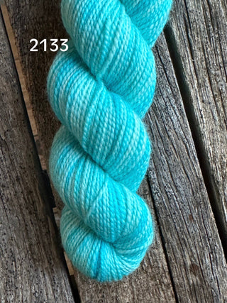 A skein of Koigu KPM & KPPPM Fingering Weight Yarn in color 2133, a hand-dyed turquoise blue and white variegated shade, rests on a rustic wooden surface.