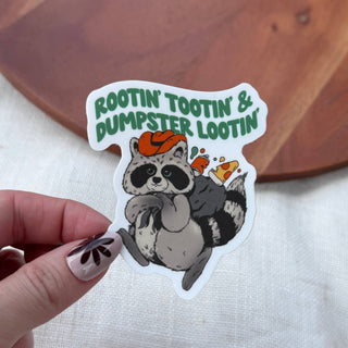 The Rootin' Tootin' & Dumpster Lootin' Raccoon Sticker shows a trash panda in an orange cowboy hat, holding its belly and giving a thumbs-up with the phrase “ROOTIN’ TOOTIN’ & DUMPSTER LOOTIN’” above.