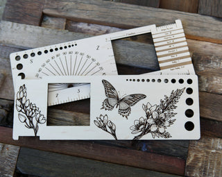 The Butterfly "Almost Everything" Ruler is a laser-cut wooden knitting gauge with floral and butterfly engravings, yarn weight guide, and measurement holes, beautifully displayed on a rustic wooden surface.