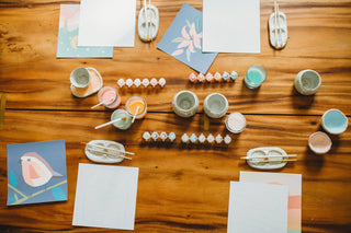 A wooden table displays the Wildflowers Meditative Art Paint by Number Kit with paint pots, brushes, water cups, and partially painted floral illustrations, all arranged for a relaxing, mindful painting session.