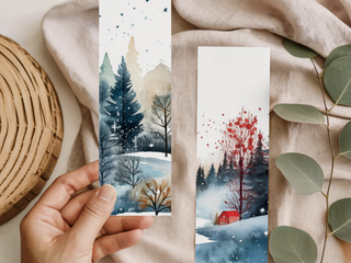 A hand holds a watercolor-painted Holiday Bookmark for Knitters with snowy trees and a cloudy sky; another bookmark nearby features trees, a red house, and snowfall, both on beige fabric with eucalyptus leaves and a wicker tray.