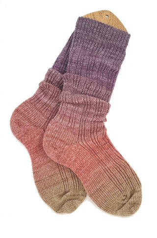 These ribbed gradient socks, crafted from Solemate Sock Yarn, transition from olive green at the toes to red and purple at the cuffs. They are displayed on a wooden sock blocker.