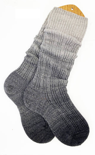 A pair of long, ribbed knit gradient socks made with soft Solemate Sock Yarn transitions from dark gray at the toes to light gray at the top, shown flat on a wooden sock blocker against a white background.
