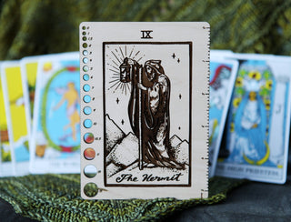 The ✨ The Hermit ✨ Tarot Knitting Needle Gauge & Ruler features a hooded figure with a staff and lantern, standing upright on a green textured surface. Other colorful tarot cards appear blurred in the background.