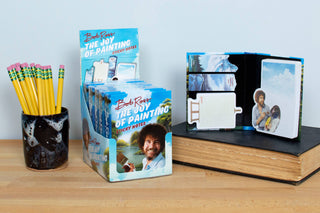 A display of Bob Ross Sticky Notes with painting designs sits on a wooden surface beside a glass of pencils and a closed book.