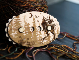 The Baba Yaga Row Counter, an oval wooden knitting accessory engraved with trees, a house, a crescent moon, and birds and featuring three numbered holes, is displayed in front of a ball of multicolored yarn and loose strands on a dark surface.