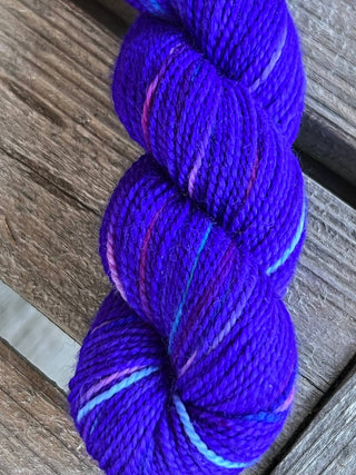 A skein from the KPM and KPPPM Variegated Color Collection in color P491—bright purple with hints of pink and blue, ideal for hand knitting—rests on a weathered wooden surface.
