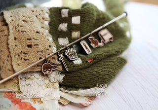 Resting on green knitted fabric, the Princess Bride Knitting Stitch Markers—featuring six enchanting charms (skull, staff, crown, sword, rabbit, and book)—add a magical touch to your project. These whimsical accessories double as stitch markers and come with a smooth maple wood knitting needle for comfortable use. Lace and patterned fabric scraps complete the charming scene.