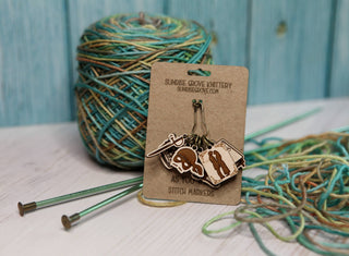 A ball of variegated yarn, knitting needles, and a card displaying quirky Princess Bride Knitting Stitch Markers made from maple wood on a wooden surface with a blue wooden background.