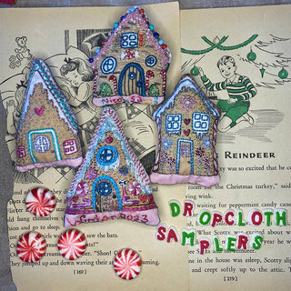 Four embroidered house-shaped ornaments and three peppermint candies from the Gingerbread Houses Embroidered Ornaments Sampler rest on open book pages, with colorful pins and stitched details adding extra holiday charm.