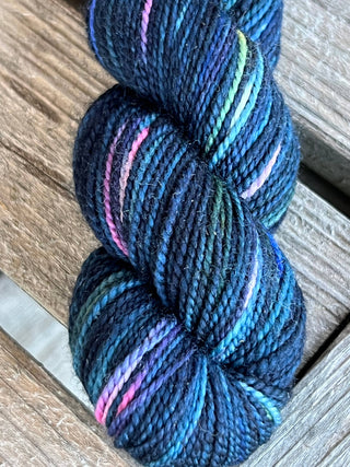 A close-up of a KPM and KPPPM Variegated Color Collection Skein in dark blue with pink, purple, green, and turquoise accents, perfect for hand knitting, resting on a wooden surface.