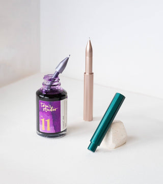 A metallic pen with its cap off is in a purple ink bottle labeled tom & midi 11. Next to it, The Wren - Refillable Writing Pen in rose gold stands upright, while a green pen cap rests on a white eraser.