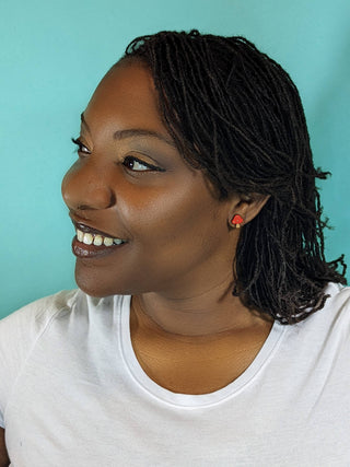 A woman with medium-dark skin and shoulder-length locs smiles while looking to the left. She wears a white shirt and handmade in the USA, hypoallergenic Mushroom Earrings, against a light blue background.