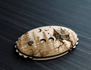 The Baba Yaga Row Counter is an oval maple wood object engraved with artwork of a house, trees, birds, and a crescent moon—adorned with three round metal accents and elegantly displayed on a dark surface.