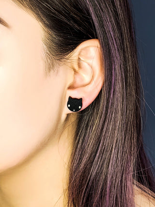 A close-up shows straight, purple-tinged hair tucked behind the ear wearing 🐈‍⬛ Black Cat Earrings. The dark background highlights these playful handmade earrings, perfect for Halloween.