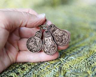 A hand holds three 💡 Terrarium Light Bulb Progress Keepers 💡—wooden earrings engraved with nature motifs like plants, leaves, and a frog. Perfect as crochet markers or knitting progress keepers against a green woven fabric background.
