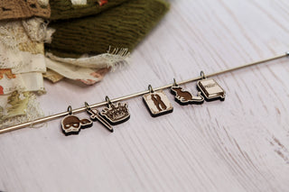 A metal rod displays the Princess Bride Knitting Stitch Markers—five charms featuring a skull, a crown with a sword, a silhouette of a person, a cat, and a book labeled “Pride.” In the background, textiles and knitting accessories rest on a white maple wood surface.