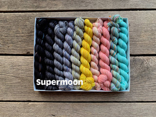 A Spooky Season Koigu Pencil Boxes set in Supermoon color, featuring ten gradient Merino wool mini skeins, sits on a wooden surface. “Supermoon” appears in bold white text at the image’s lower left.