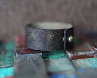 The Bee and Moon Leather Ruler Cuff, a dark brown reclaimed leather bracelet with a worn texture and round brass button, rests on a multicolored wooden surface.