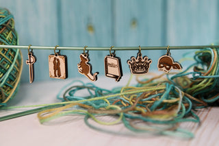 Six Princess Bride Knitting Stitch Markers, each shaped like a sword, person, rat, book, crown, and mask, hang from a knitting needle. Crafted from maple wood, these unique accessories are beautifully displayed with colorful yarn in the background and foreground on a light wooden surface.