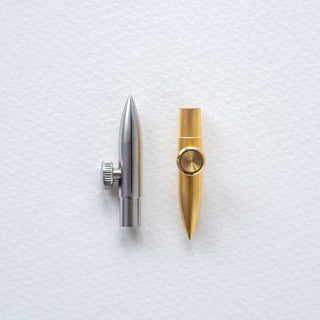 Two metallic One-Dip-Nib calligraphy nibs—one silver, one gold—with side knobs for adjustable line variation stand upright on a white textured surface, their bullet shapes evoking elegance and precision.