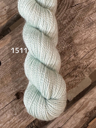 A skein of pale mint green Koigu KPM & KPPPM Fingering Weight Yarn, labeled 1511, is displayed on a rustic wooden surface—perfect for knitting or crochet projects.