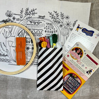 The Original Embroidery Sampler Kit includes printed fabric, a wooden hoop, orange felt, assorted threads, a striped bag, and two instruction cards for an easy tutorial—designed by Rebecca Ringquist and shown on a gray surface.