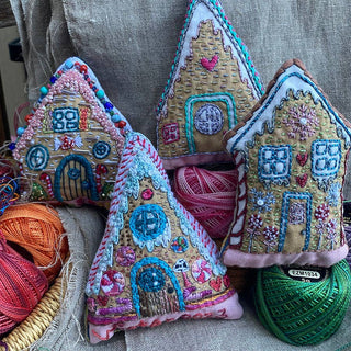 The Gingerbread Houses Embroidered Ornaments Sampler features four house-shaped fabric ornaments adorned with colorful threads, beads, and intricate patterns, displayed on textured cloth with vibrant embroidery floss.