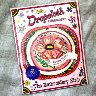 A vibrant Cosmic Poppy Embroidery Kit package labeled “Dropcloth Embroidery” displays a finished “Cosmic Poppy” floral sampler in the center, framed by decorative accents and playful patterns on a pink background.