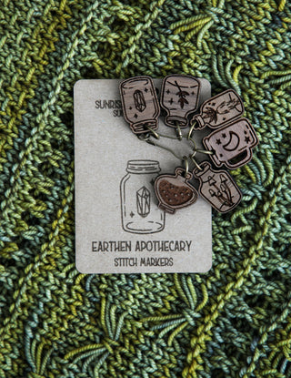 The Apothecary Bottle Stitch Marker Set features walnut wood markers with jar and cup designs, displayed on textured green and blue knitted fabric—charming accessories to enhance your knitting projects.