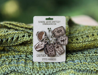 The Apothecary Bottle Stitch Marker Set features walnut wood markers with nature and celestial motifs, displayed on a branded card atop green and yellow knitted fabric—unique accessories for your project bag.