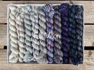 A Spooky Season Koigu Pencil Boxes set in the “Full Moon” colorway features twelve mini skeins of Merino wool yarn, ranging from pale gray and pastel to deep navy and black, displayed on a wooden surface.