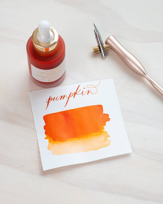 A card with orange watercolor swatches and “pumpkin” in cursive rests on a light surface beside a calligraphy pen and a bottle of Premium Calligraphy Ink, featuring a gold cap and dropper.