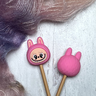 Knitting Needle Stitch Stoppers: Two wooden sticks with pink, bunny-shaped stoppers—one displays a smiling face with big eyes, the other shows the back—set against pastel textured fabric on a white surface.