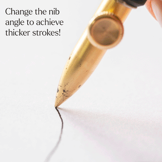 A close-up of the One-Dip-Nib gold fountain pen with a calligraphy nib writing a bold black line on white paper, with text: "Change the nib angle for thicker strokes and explore line variation!.