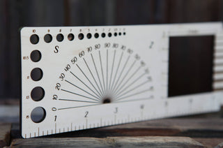 A close-up of the Luna Moth “Almost Everything” Ruler, a wooden fiber arts tool with circular cutouts, a protractor, bottom-edge ruler, and rectangular cutout on the right, resting on a wooden surface.