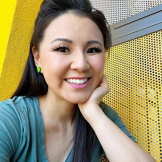 A woman with long dark hair and a teal shirt smiles at the camera, resting her chin on her hand. She wears Alien Joyride Earrings while sitting in front of a yellow wall and a perforated metal screen.