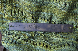 The Bee and Moon Leather Ruler Cuff features engraved designs—crescent moon, spider, and faint ruler markings—on worn dark reclaimed leather, displayed atop a green textured knit with lacy patterns.