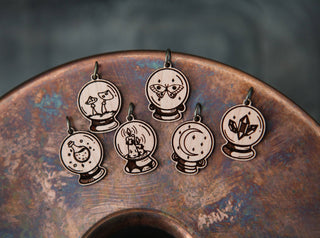 The 🔮 Crystal Ball Stitch Marker Set 🔮 features five cherry wood markers shaped like crystal balls, engraved with mystical designs—a cat, moth, moon, candle, stars & crystals—perfect witchy knitting accessories on a circular metallic display.