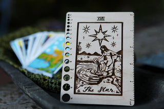 The Maple Wood Tarot Card Knitting Needle Gauge & 5 Ruler, crafted from maple, features The Star card—showing a woman by a pond beneath a large star with mountains behind. Blurred colorful tarot cards sit on a tray in the background.