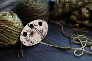 Three yarn balls and the Yarn Management Tool for Knitters, a wooden separator engraved with bee, moon, and floral designs and threaded with strands through its holes. This delightful crochet accessory is displayed on a dark, textured surface.