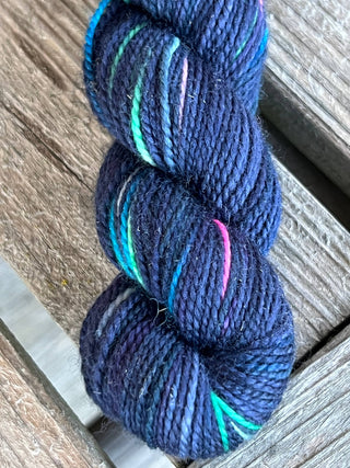 A close-up of a KPM and KPPPM Variegated Color Collection Skein in dark blue with green, turquoise, pink, and white streaks, perfect for hand knitting, displayed on a weathered wooden surface.